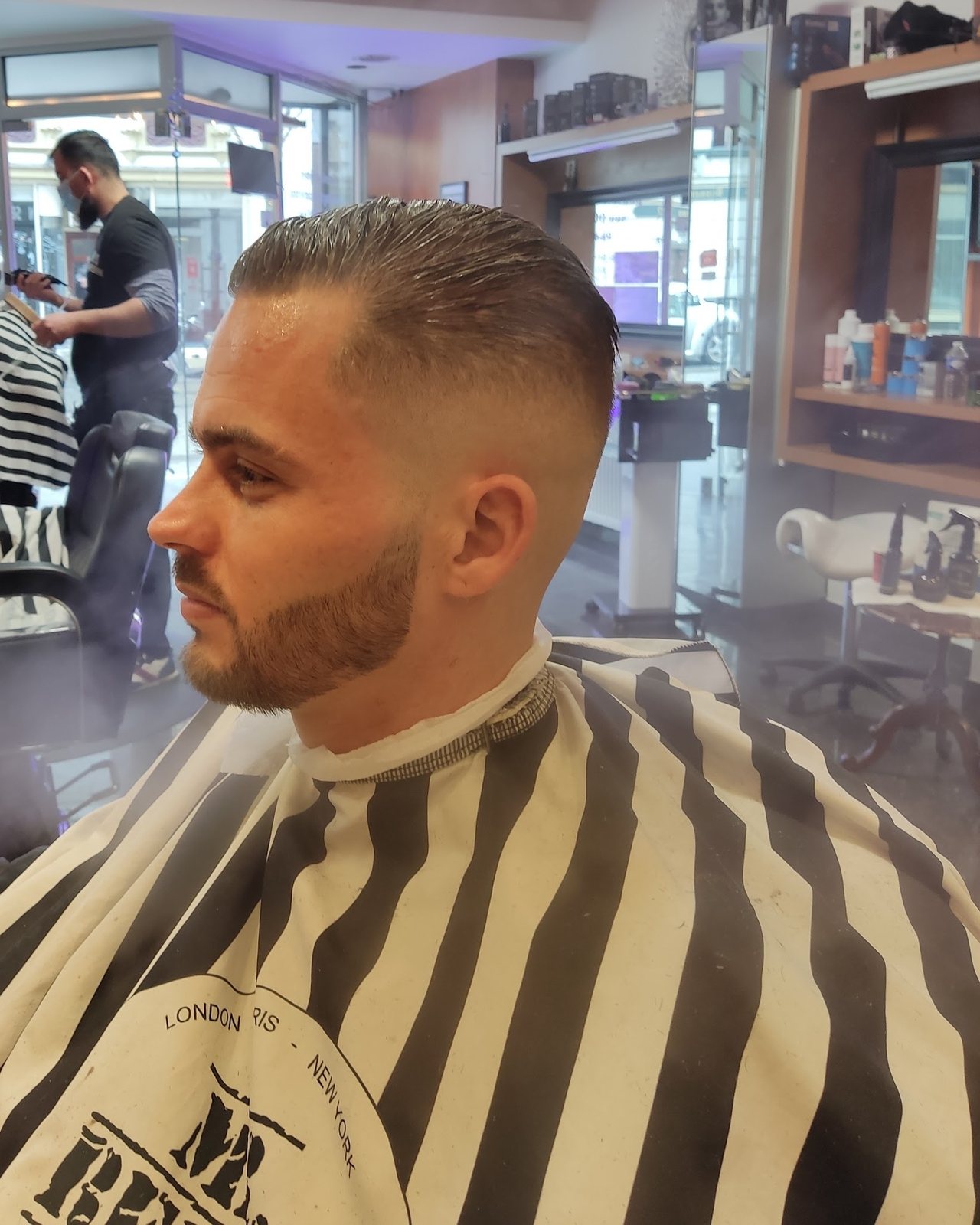 Klant in de stoel met een verse skin fade en strak getrimde baard, op de achtergrond knipt een collega